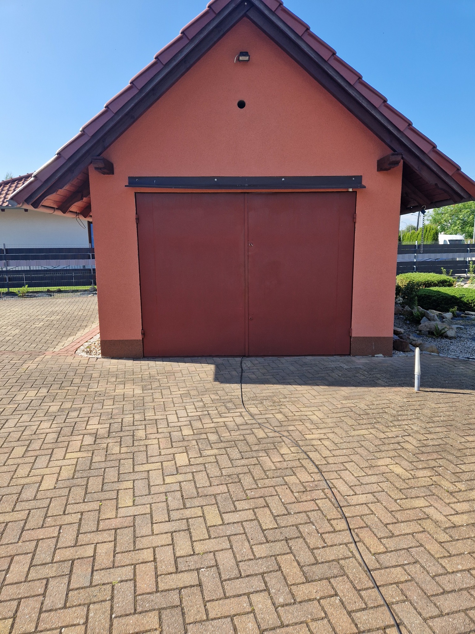 Czerwona brama garażowa na tle kostki brukowej ułożonej w jodełkę, widoczny przewód ciśnieniowy sugerujący prace porządkowe.