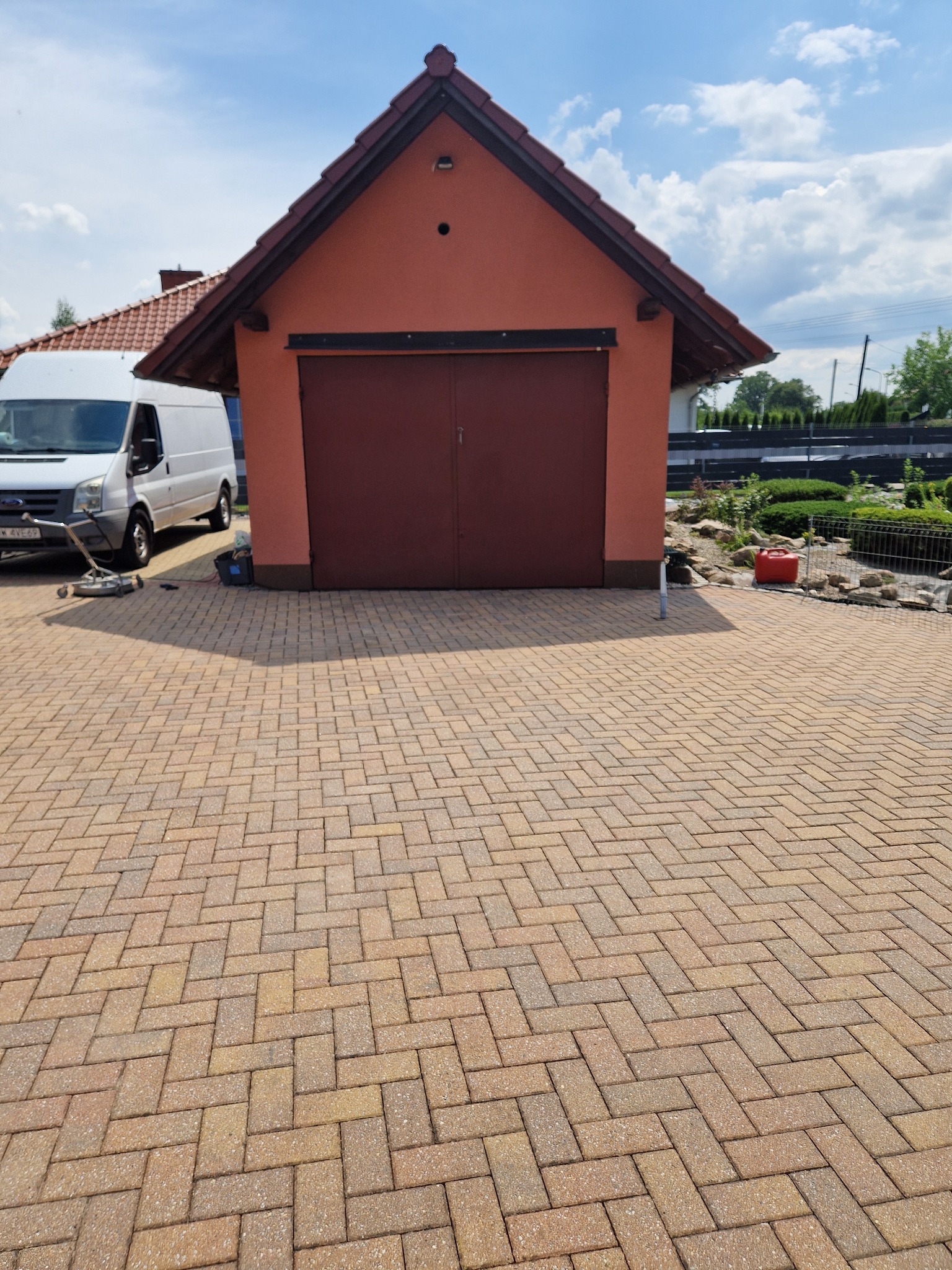 Umyta kostka brukowa ułożona we wzór jodełki, widoczny budynek gospodarczy z brązowymi drzwiami, biały van i myjka ciśnieniowa na pierwszym planie.