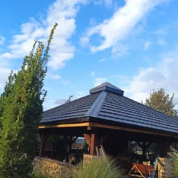 Zadaszenie altany z ciemną dachówką w ogrodzie, widok z dołu na tle błękitnego nieba z chmurami.