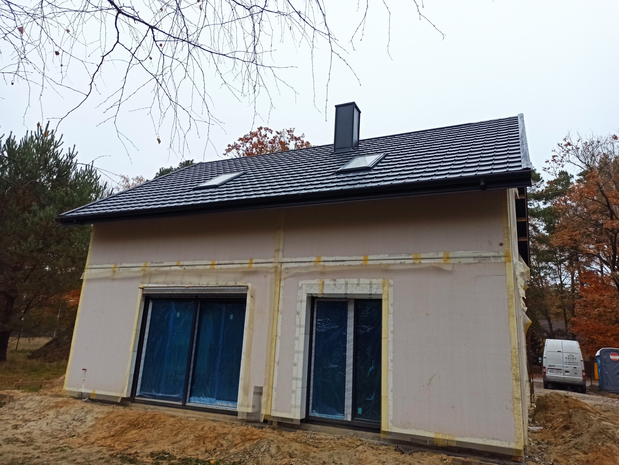 Nowy, czarny dach z dwoma oknami połaciowymi i kominem na surowym budynku w trakcie budowy, widoczne zabezpieczone okna folią, ocieplenie ścian i samochód dostawczy w tle.