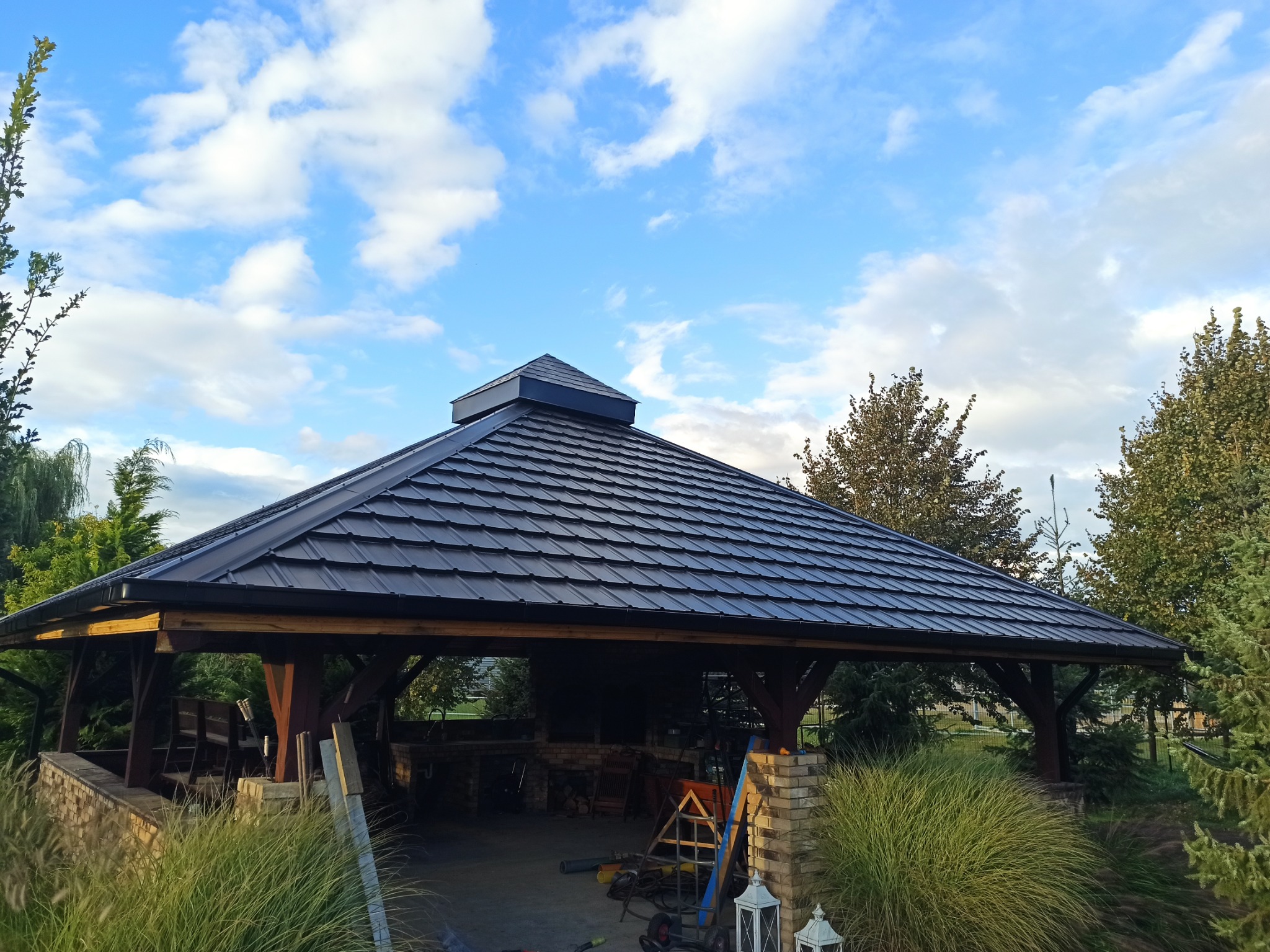Ciemna, metalowa dachówka na drewnianej altanie w ogrodzie, widok z dołu na tle błękitnego nieba z chmurami.