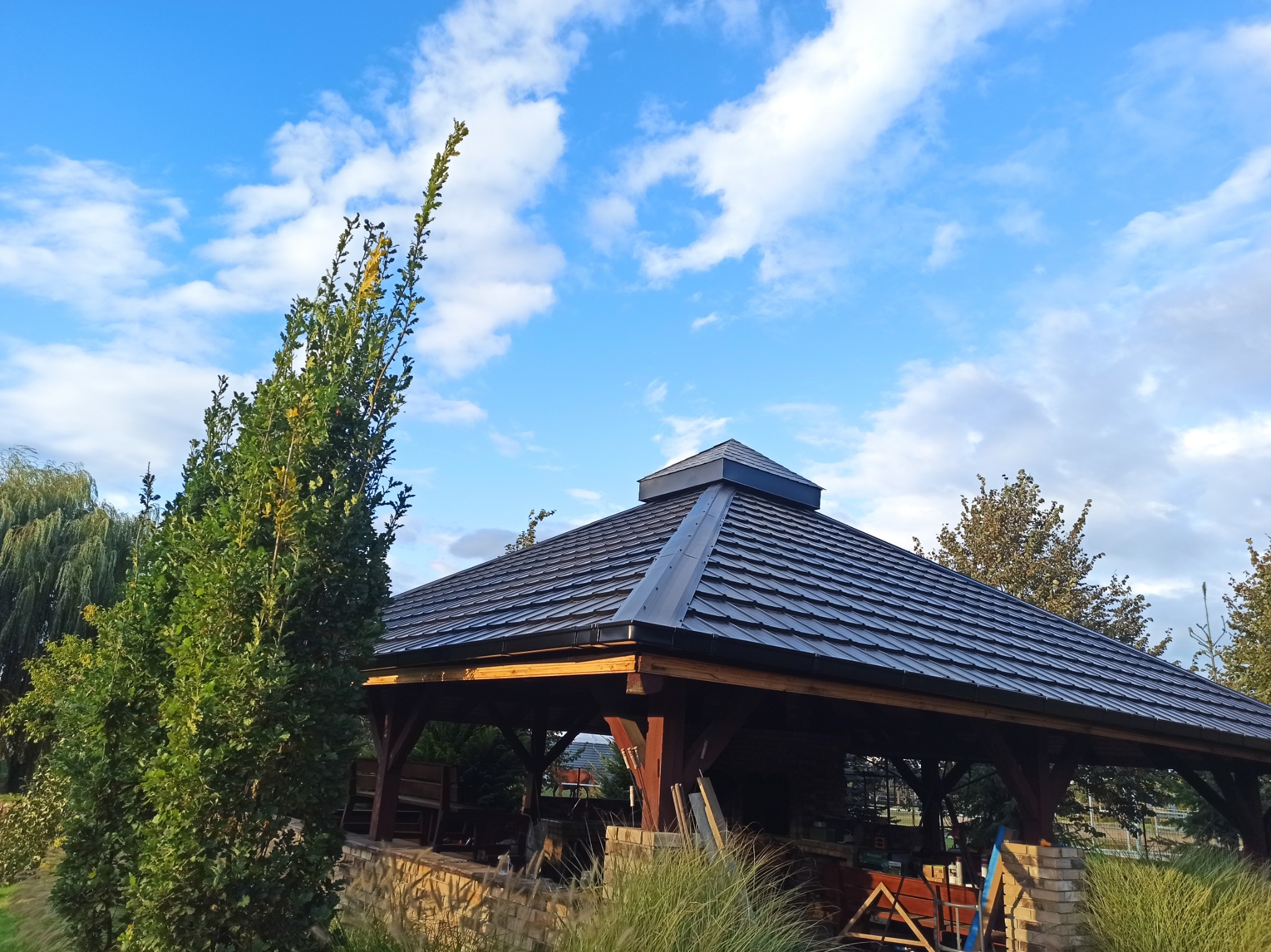 Zadaszenie altany z ciemną dachówką w ogrodzie, widok z dołu na tle błękitnego nieba z chmurami.