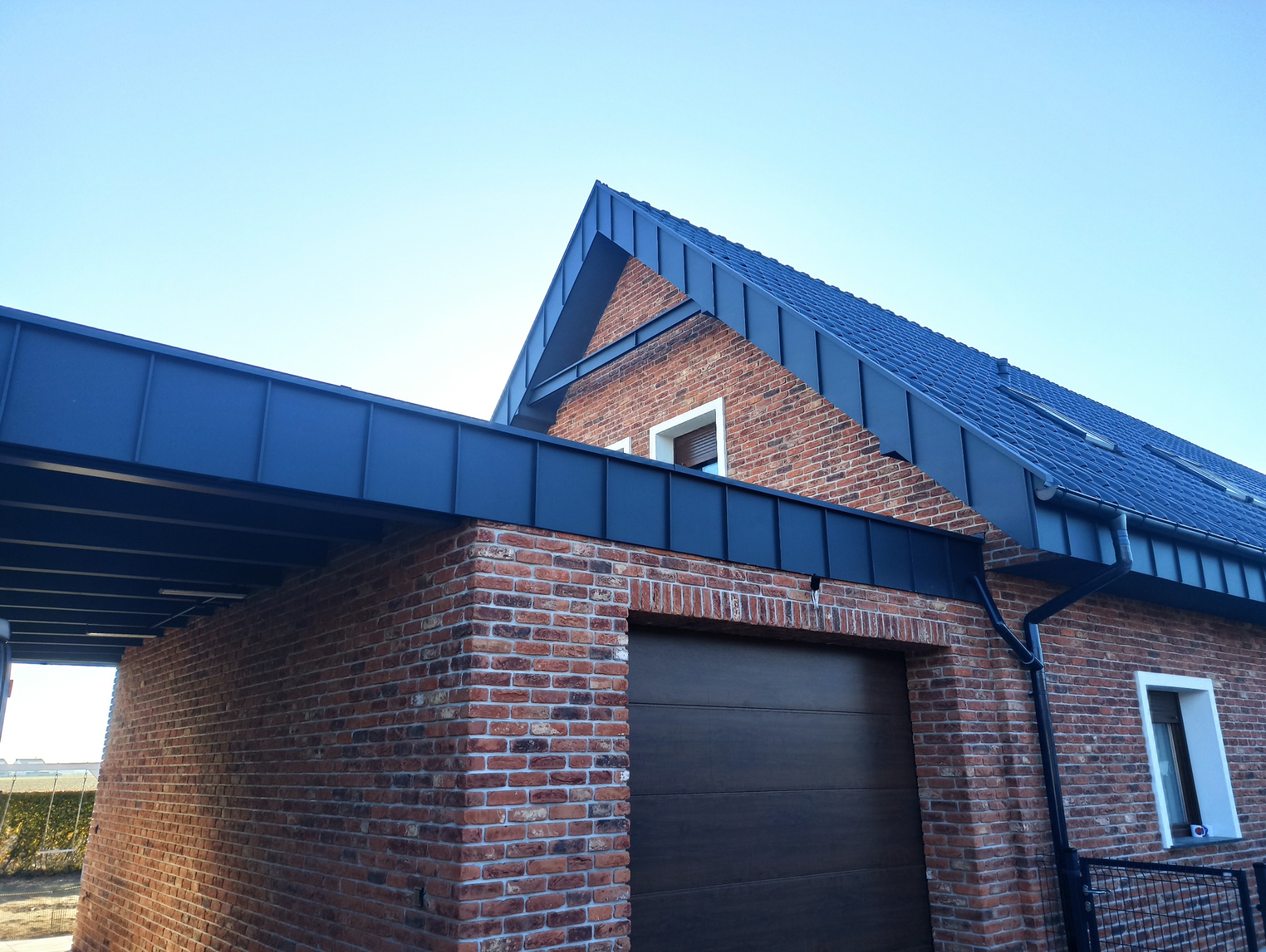 Dom z czerwonej cegły z ciemnym dachem i rynnami. Widoczny fragment garażu i zadaszenia. Ujęcie z dołu, podkreślające geometrię budynku i detale wykończenia.