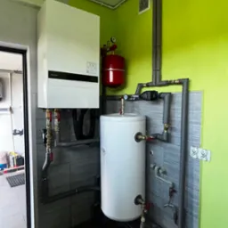 Nowoczesny, biały piec gazowy wiszący na szarej ścianie z płytkami, obok czerwony zbiornik wyrównawczy i biały zasobnik ciepłej wody, widoczne rury i zawory, ściana pomalowana na jasnozielono.