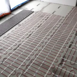 Rozłożone maty grzewcze pod panele podłogowe, widoczny fragment kratki wentylacyjnej przy oknie.