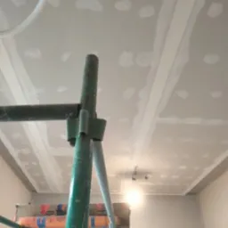 Sufit z płyt kartonowo-gipsowych w trakcie wykańczania, widoczne łączenia i miejsca po wkrętach, oświetlenie tymczasowe, fragment rusztowania malarskiego z pomarańczowymi rolkami folii ochronnej.