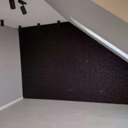 Pokój na poddaszu z czarną ścianą z cegły, szarą ścianą i jasną podłogą, oświetlenie punktowe na suficie.