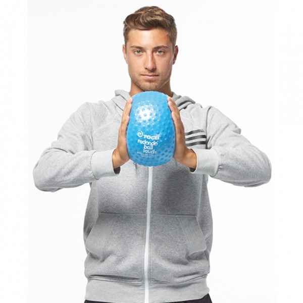 Młody mężczyzna w szarej bluzie z kapturem trzymający oburącz niebieską piłkę rehabilitacyjną z wypustkami i napisem 'TOGU redondo ball touch' na białym tle.