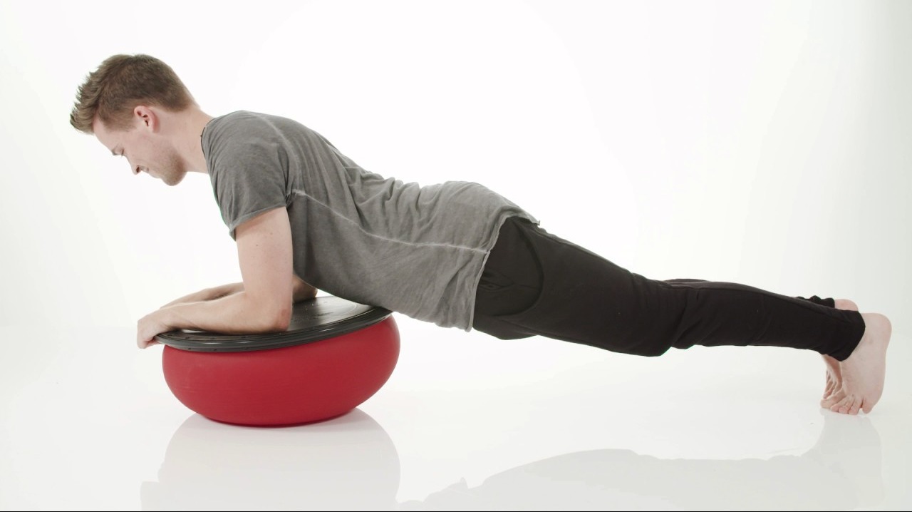 Mężczyzna w szarej koszulce i czarnych legginsach wykonuje ćwiczenie plank na czerwonej piłce Bosu na białym tle.