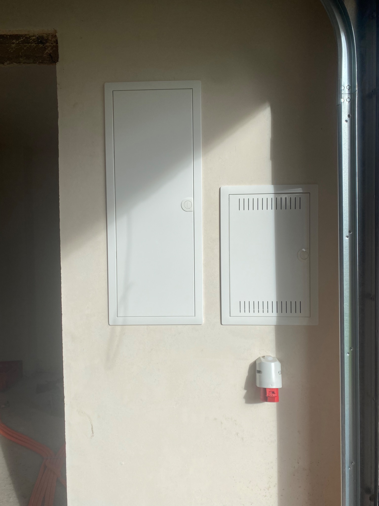 Dwie białe skrzynki elektryczne, jedna z wentylacją, zamontowane na jasnej ścianie w surowym wnętrzu, obok widoczny element bramy garażowej i gniazdo siłowe.