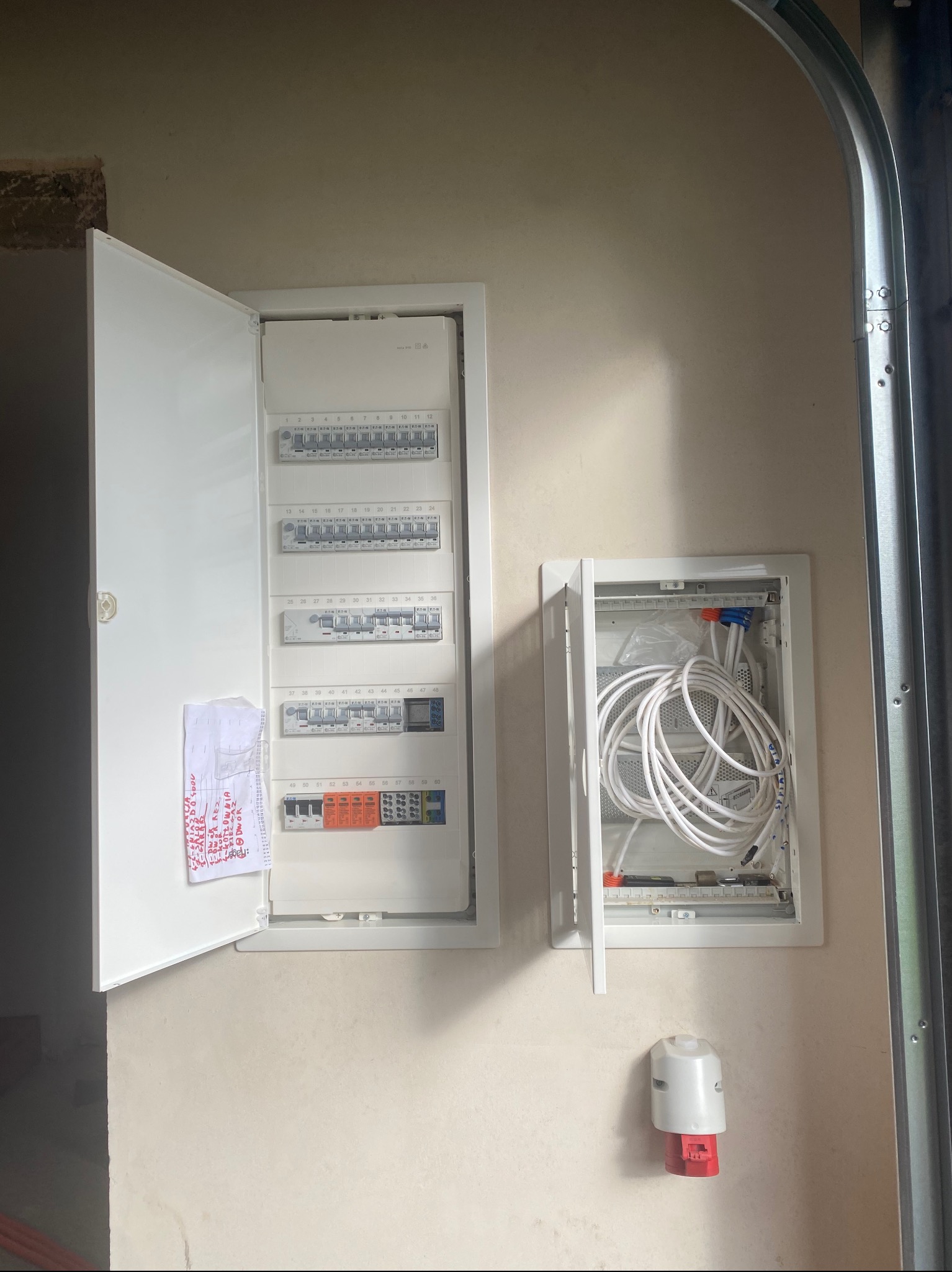 Otwarta skrzynka rozdzielcza z wyłącznikami i bezpiecznikami, notatka z odręcznymi zapiskami na drzwiach, obok druga skrzynka z przewodem światłowodowym, gniazdo siłowe na ścianie.