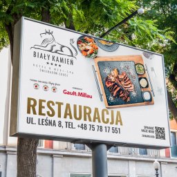 BR Studio Graficzne Barbara Rutkowska - Billboard reklamowy hotelu i restauracji Biały Kamień w Świeradowie-Zdroju, prezentujący logo, dania z menu oraz nagrody i wyróżnienia, umieszczony na tle drzew i budynku.