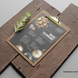 BR Studio Graficzne Barbara Rutkowska - Eleganckie menu restauracji 'Biały Kamień' na desce z klipsem, prezentujące sezonowe dania, takie jak tatar z jelenia, grillowany ser kozi i banoffee pie, z gałązką rozmarynu w tle.
