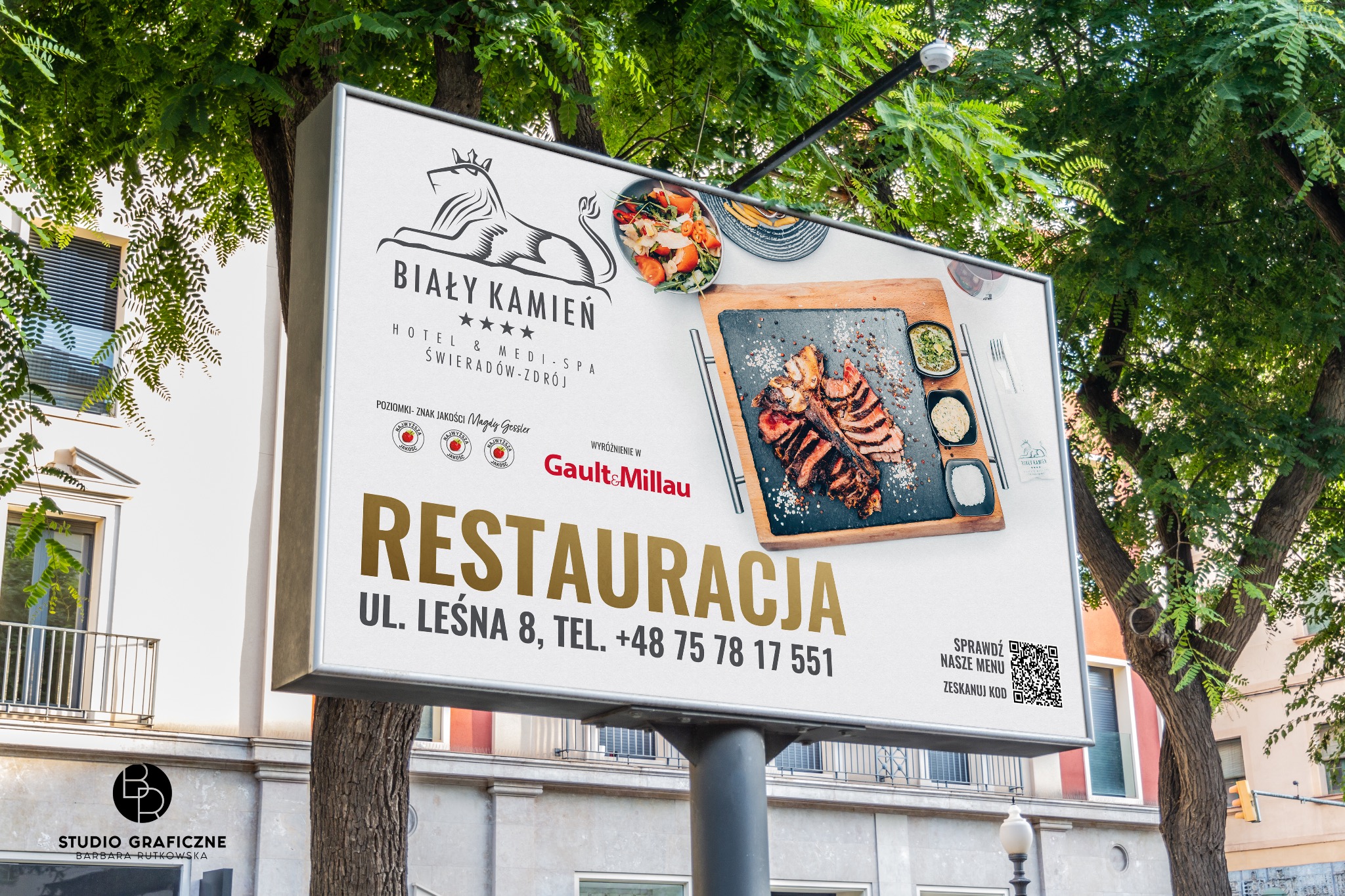 Billboard reklamowy hotelu i restauracji Biały Kamień w Świeradowie-Zdroju, prezentujący logo, dania z menu oraz nagrody i wyróżnienia, umieszczony na tle drzew i budynku.
