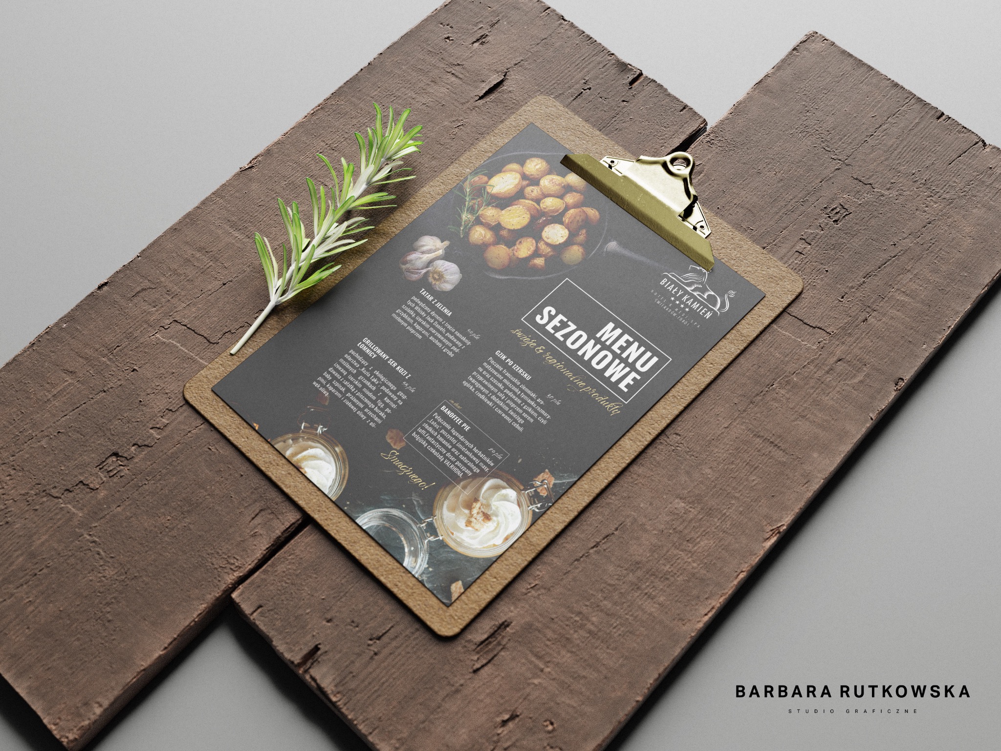 Eleganckie menu restauracji 'Biały Kamień' na desce z klipsem, prezentujące sezonowe dania, takie jak tatar z jelenia, grillowany ser kozi i banoffee pie, z gałązką rozmarynu w tle.