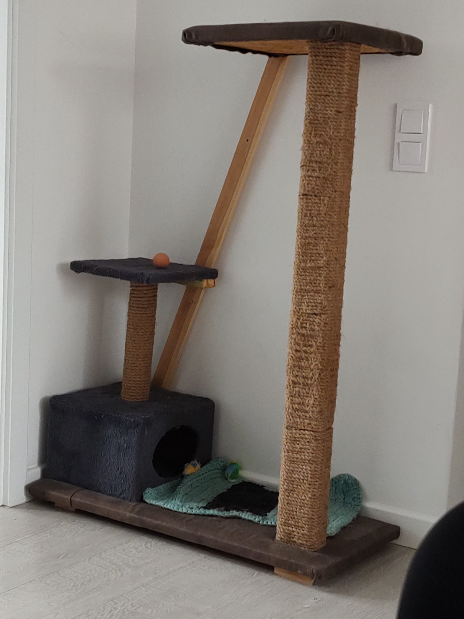 Wielopoziomowy drapak dla kota z tunelem, platformami obitymi ciemną tkaniną i słupkami owiniętymi sizalem, ustawiony w rogu pokoju na jasnej podłodze.