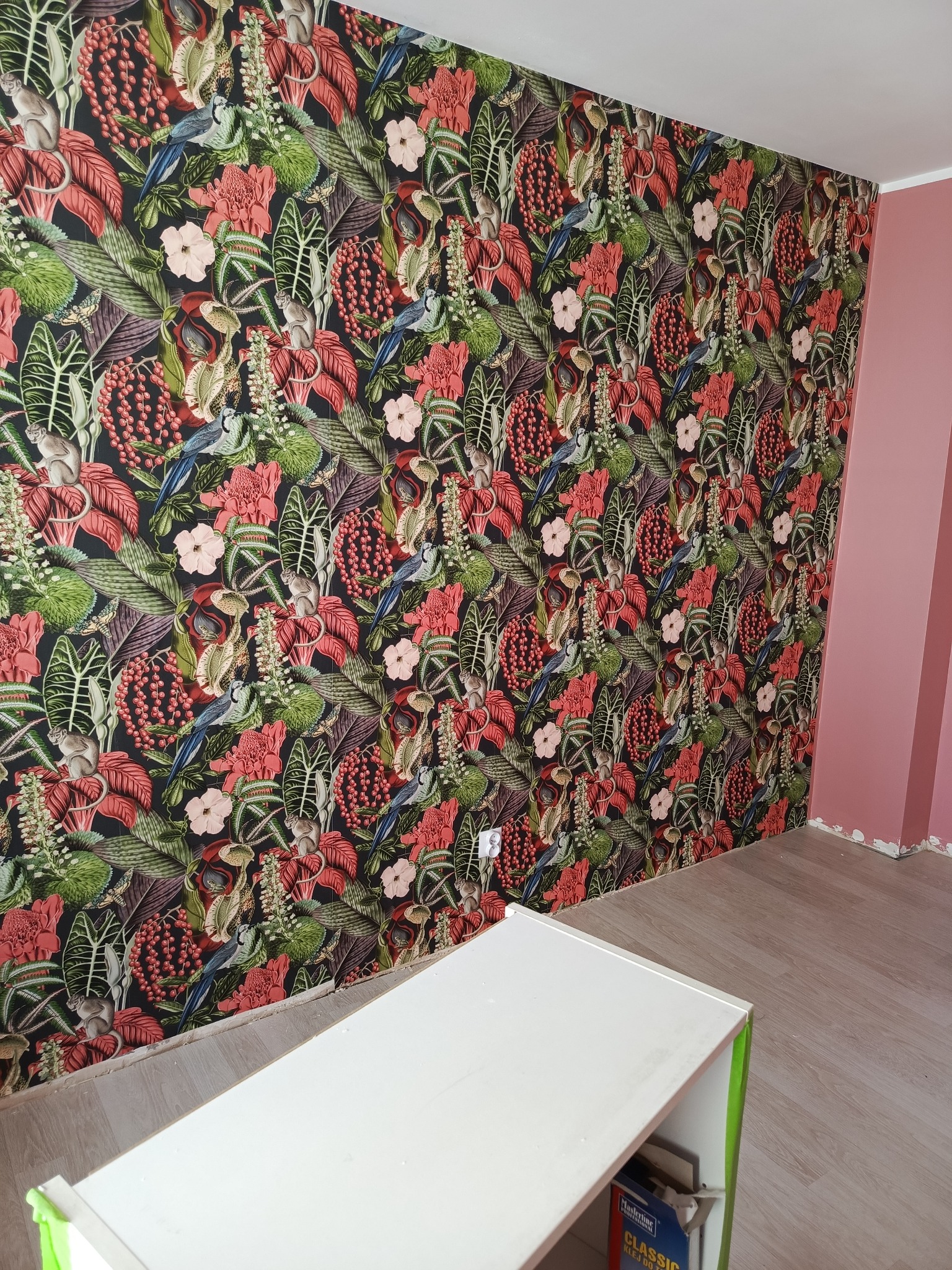 Ściana wytapetowana w tropikalny wzór z motywami roślinnymi, ptaków i małp, z fragmentem różowej ściany i białym meblem w dolnej części kadru.