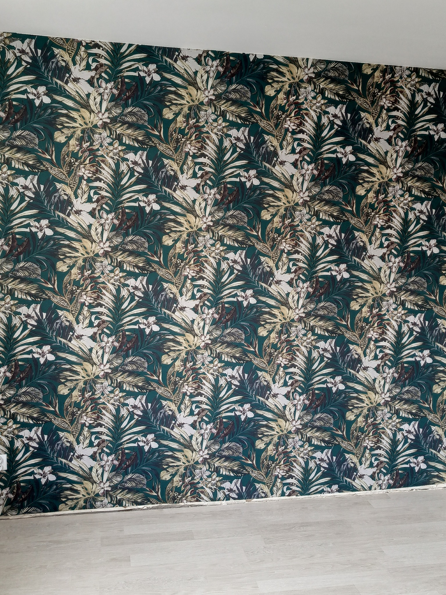 Ściana pokryta tapetą w tropikalne liście i kwiaty, z widocznym fragmentem jasnej podłogi i gniazdkiem elektrycznym po lewej stronie.