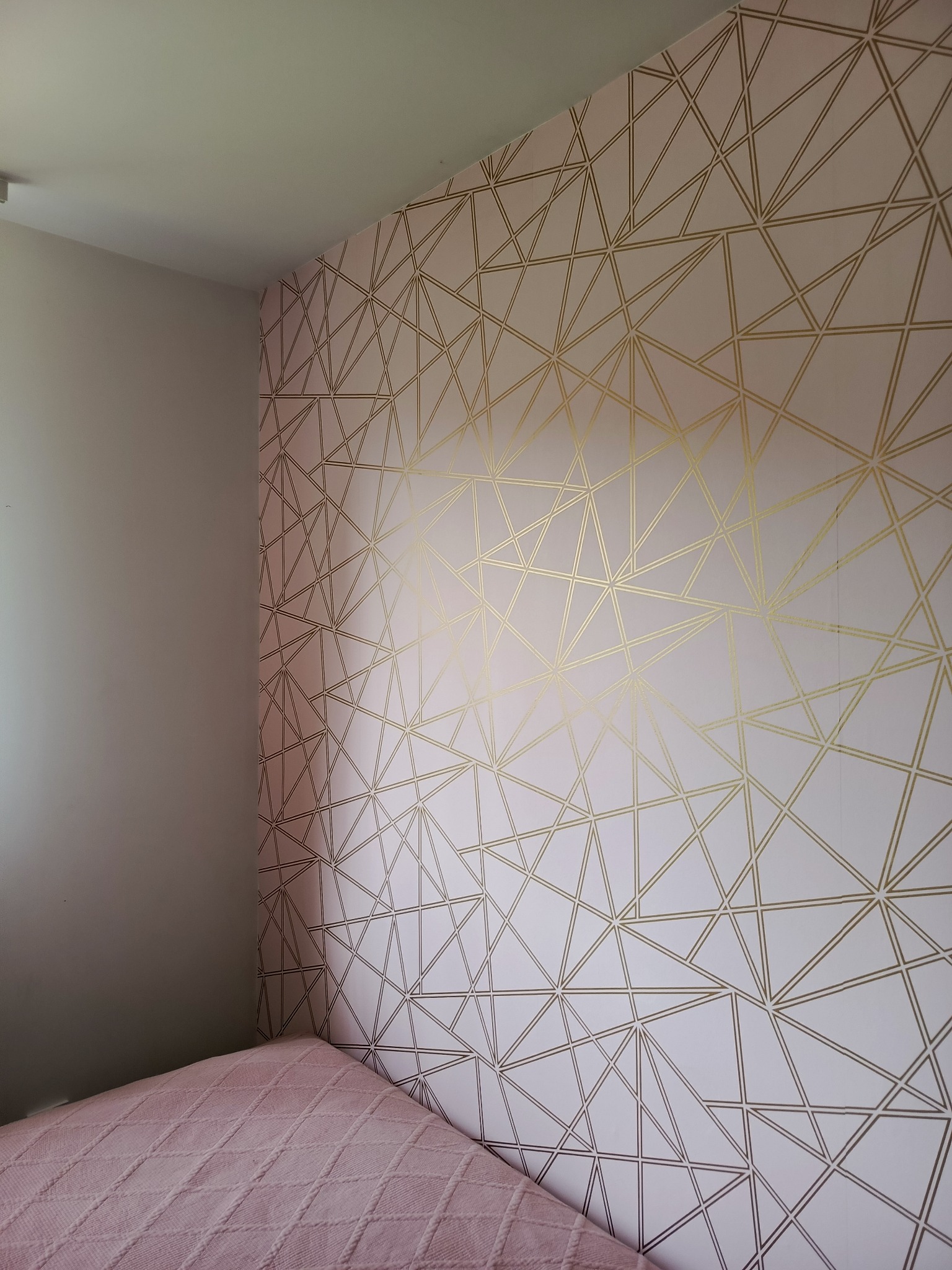 Różowa ściana z geometrycznym wzorem złotych linii w sypialni, widoczny fragment łóżka z pikowaną narzutą w tym samym odcieniu.