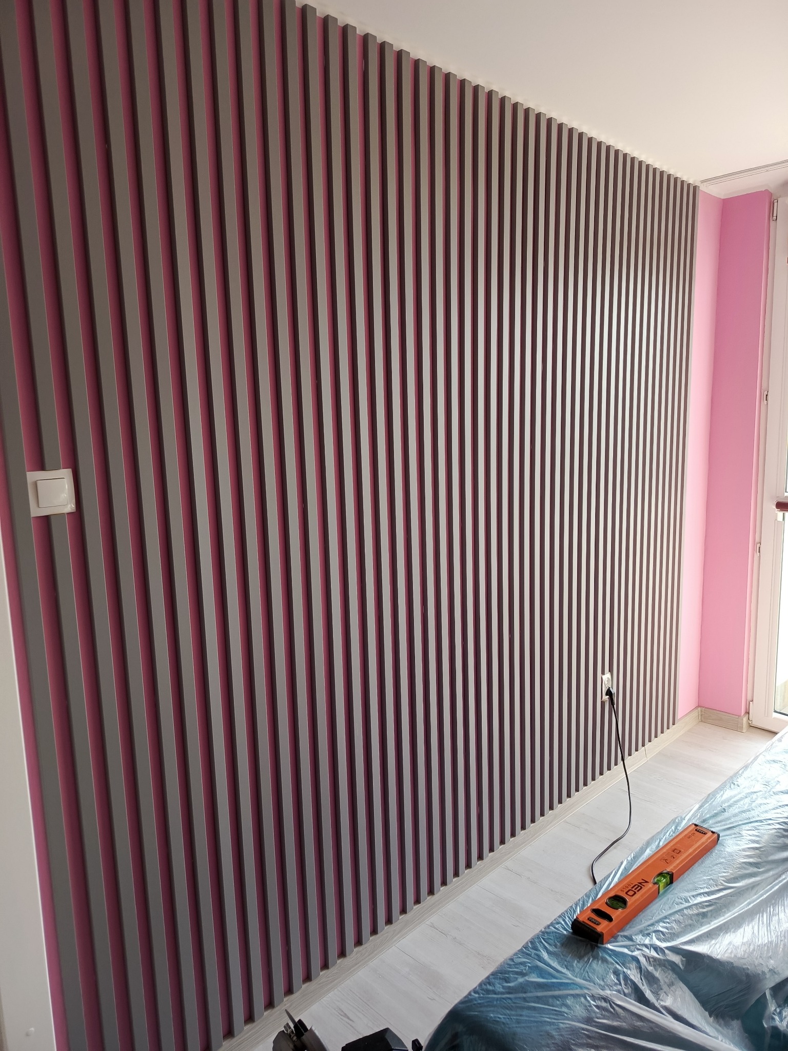 Ściana z szarymi pionowymi panelami dekoracyjnymi z różowym tłem, widoczny poziomica, zabezpieczona folią podłoga i gniazdko elektryczne.