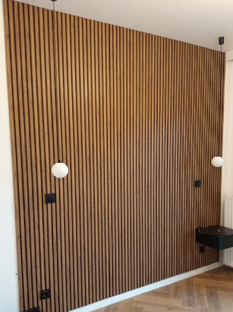 Ściana wykończona panelami drewnianymi z czarnymi szczelinami, oświetlona kulistymi lampami wiszącymi i z zamontowaną czarną szafką nocną.