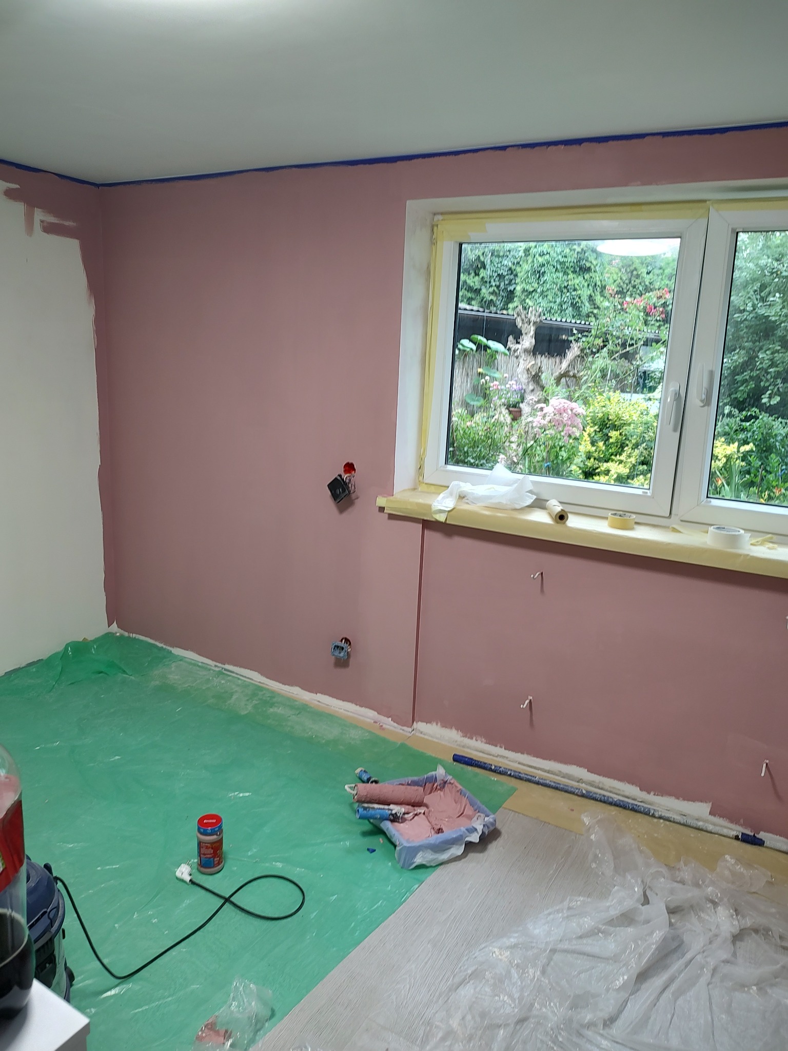 Pokój w trakcie odświeżania: ściany pomalowane na różowo, zabezpieczone okno taśmą malarską, podłoga przykryta folią ochronną, widoczny wałek malarski z resztkami farby i puszka farby na podłodze.