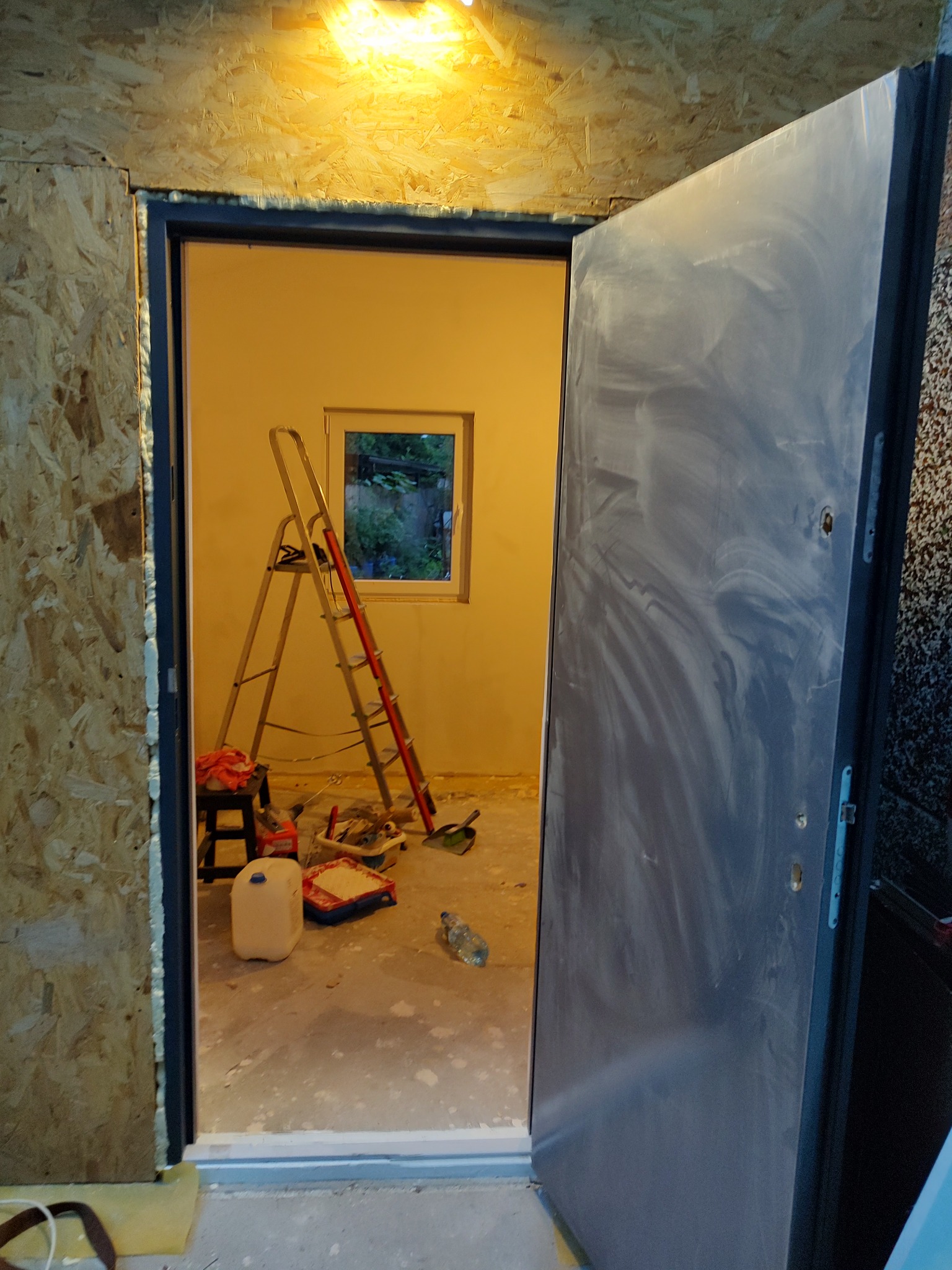 Wnętrze pomieszczenia w trakcie remontu z otwartymi drzwiami, widoczna drabina oparta o ścianę pod oknem, narzędzia malarskie, kanister z farbą i butelka wody na podłodze, ściany przygotowane...