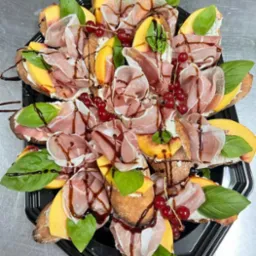 Apetyczna kompozycja kanapek na czarnym talerzu: pieczywo z kremowym serem, brzoskwiniami, szynką parmeńską, bazylią, czerwoną porzeczką i sosem balsamicznym.