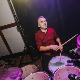 Zespół muzyczny Simon - Perkusista w bordowej koszuli, z pałeczkami w dłoniach, przygotowuje się do gry na perkusji w oświetleniu scenicznym.