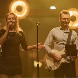 Wokalistka w czarnej sukience śpiewa do mikrofonu, gitarzysta gra na białej gitarze elektrycznej, w tle klawiszowiec i ciepłe światła sceniczne.