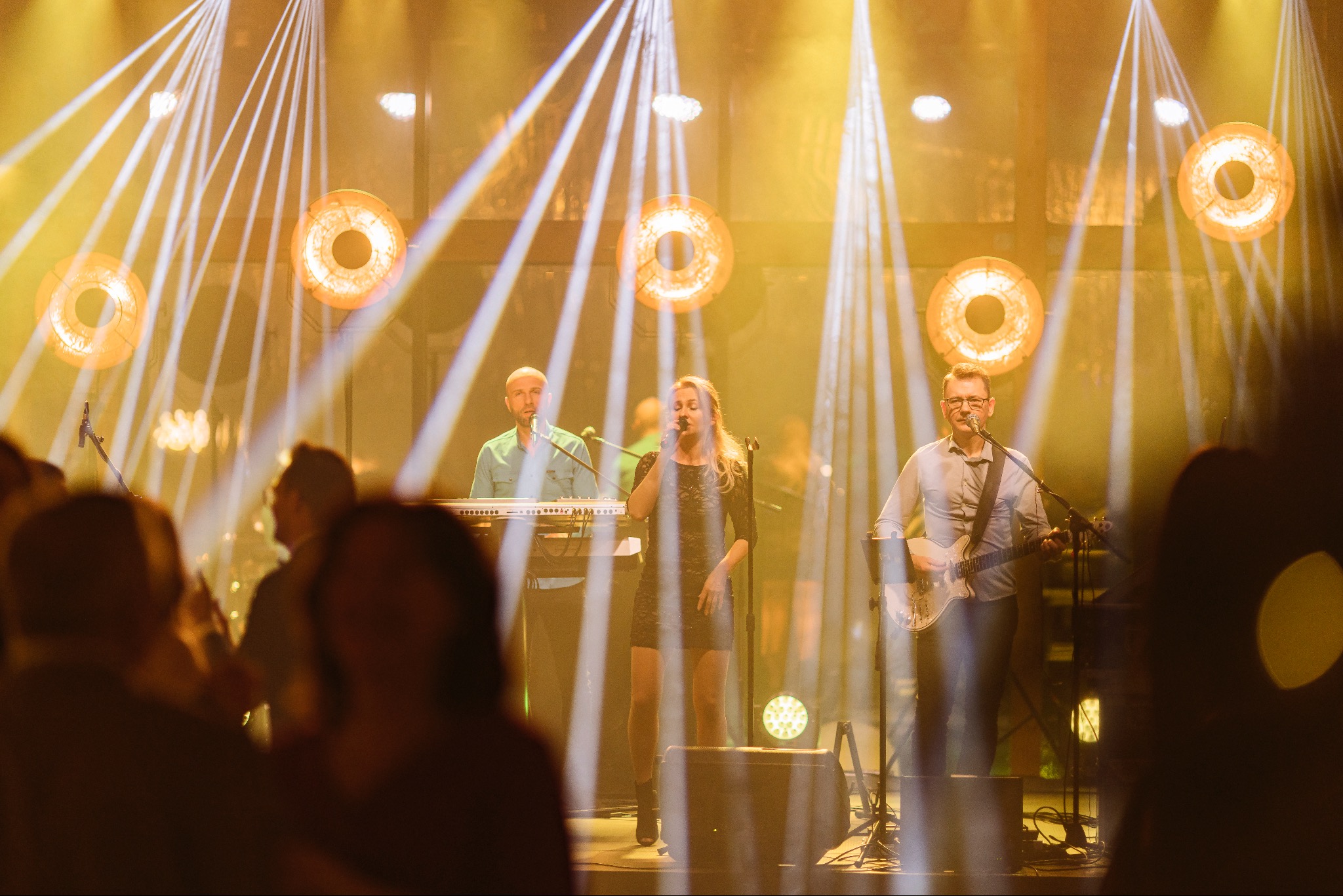 Zespół muzyczny na scenie podczas występu, oświetlony reflektorami, wokalistka w czarnej sukience śpiewa do mikrofonu, obok klawiszowiec i gitarzysta, w tle widoczna publiczność.