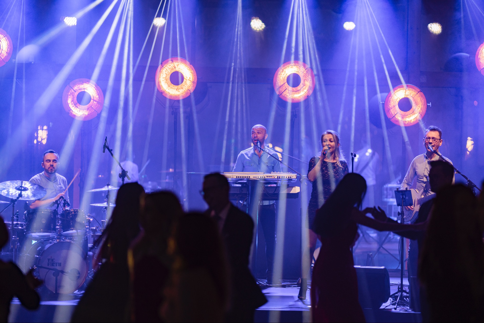 Zespół grający na scenie z oświetleniem punktowym, widoczny perkusista, klawiszowiec, wokalistka i gitarzysta, w tle dekoracyjne okrągłe lampy, w pierwszym planie rozmazane sylwetki tańczących osób.
