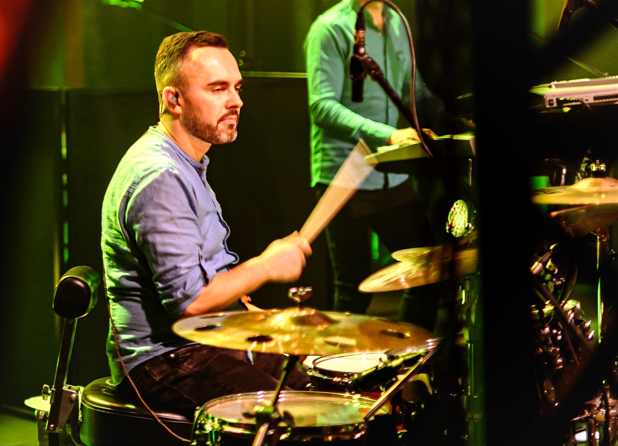 Perkusista grający na perkusji podczas występu na scenie, w tle klawiszowiec, oświetlenie sceniczne w odcieniach zieleni i żółci.