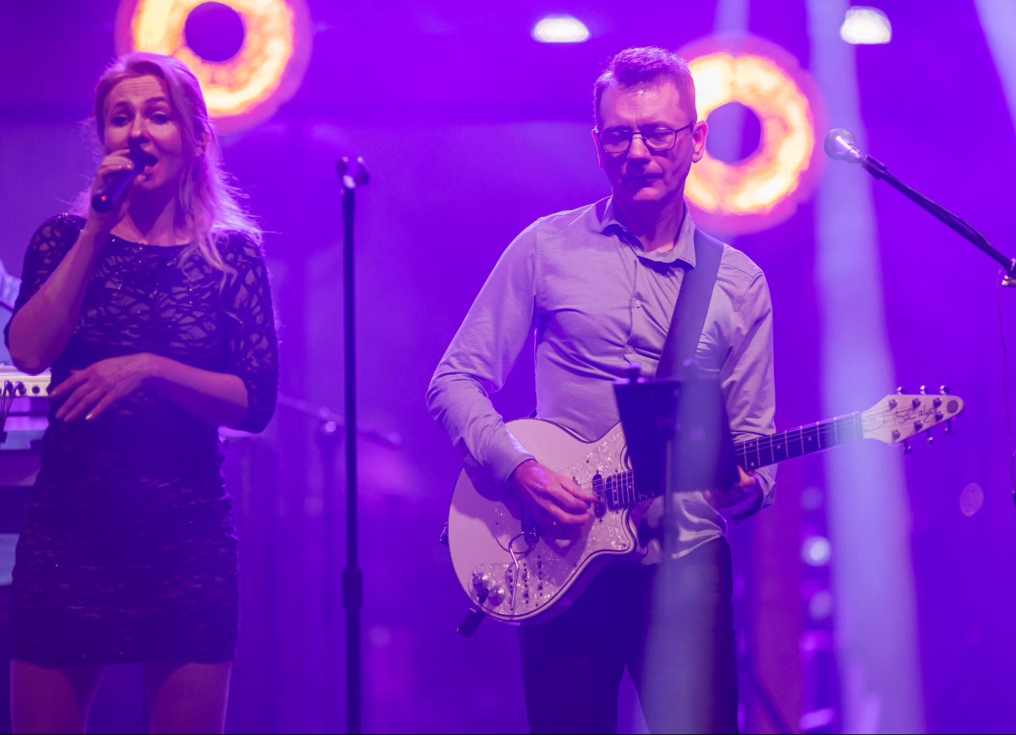 Wokalistka z mikrofonem i gitarzysta z białą gitarą elektryczną na scenie oświetlonej fioletowym światłem, z dekoracyjnymi okrągłymi lampami w tle.