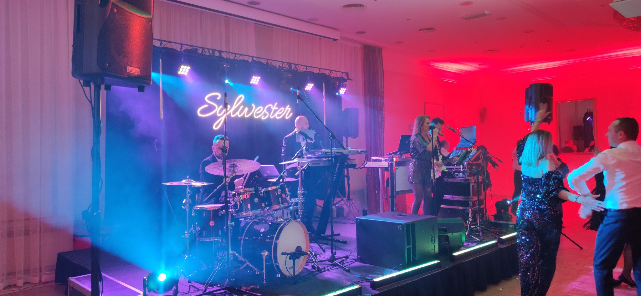 Zespół muzyczny grający na scenie podczas imprezy Sylwestrowej, oświetlonej niebieskim i czerwonym światłem, z widocznymi instrumentami: perkusja, keyboard oraz wokalistka z mikrofonem, przed sceną...