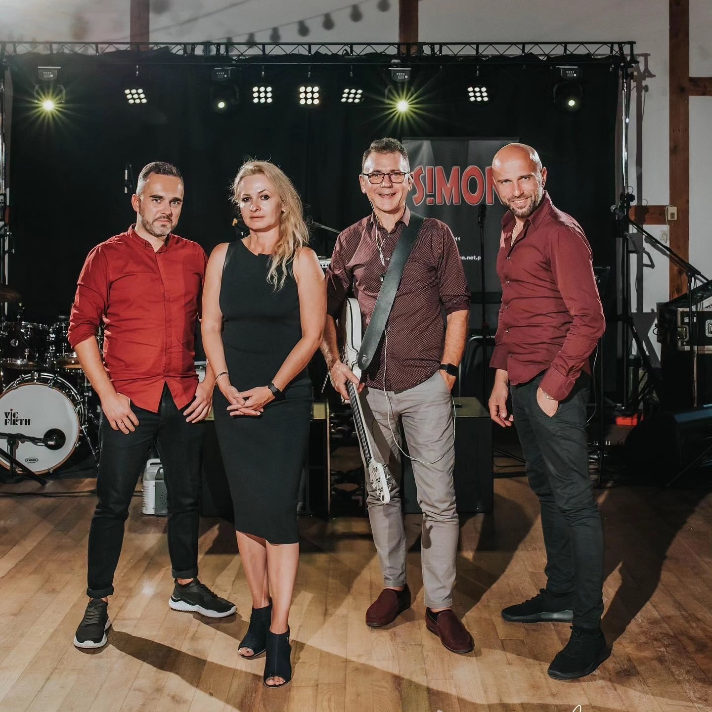 Portret zespołu muzycznego na scenie przed koncertem, z logo 'SIMON' widocznym w tle, oświetlonymi reflektorami i perkusją po lewej stronie.
