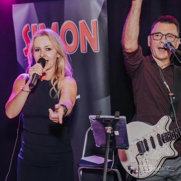 Wokalistka w czarnej sukience wskazująca palcem i gitarzysta w okularach z podniesioną ręką, występujący na scenie z oświetleniem punktowym i banerem w tle.