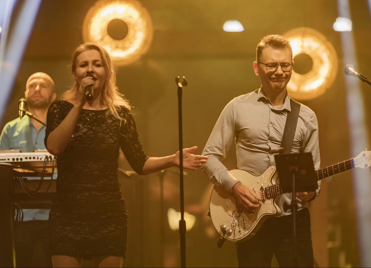 Wokalistka w czarnej sukience śpiewa do mikrofonu, gitarzysta gra na białej gitarze elektrycznej, w tle klawiszowiec i ciepłe światła sceniczne.