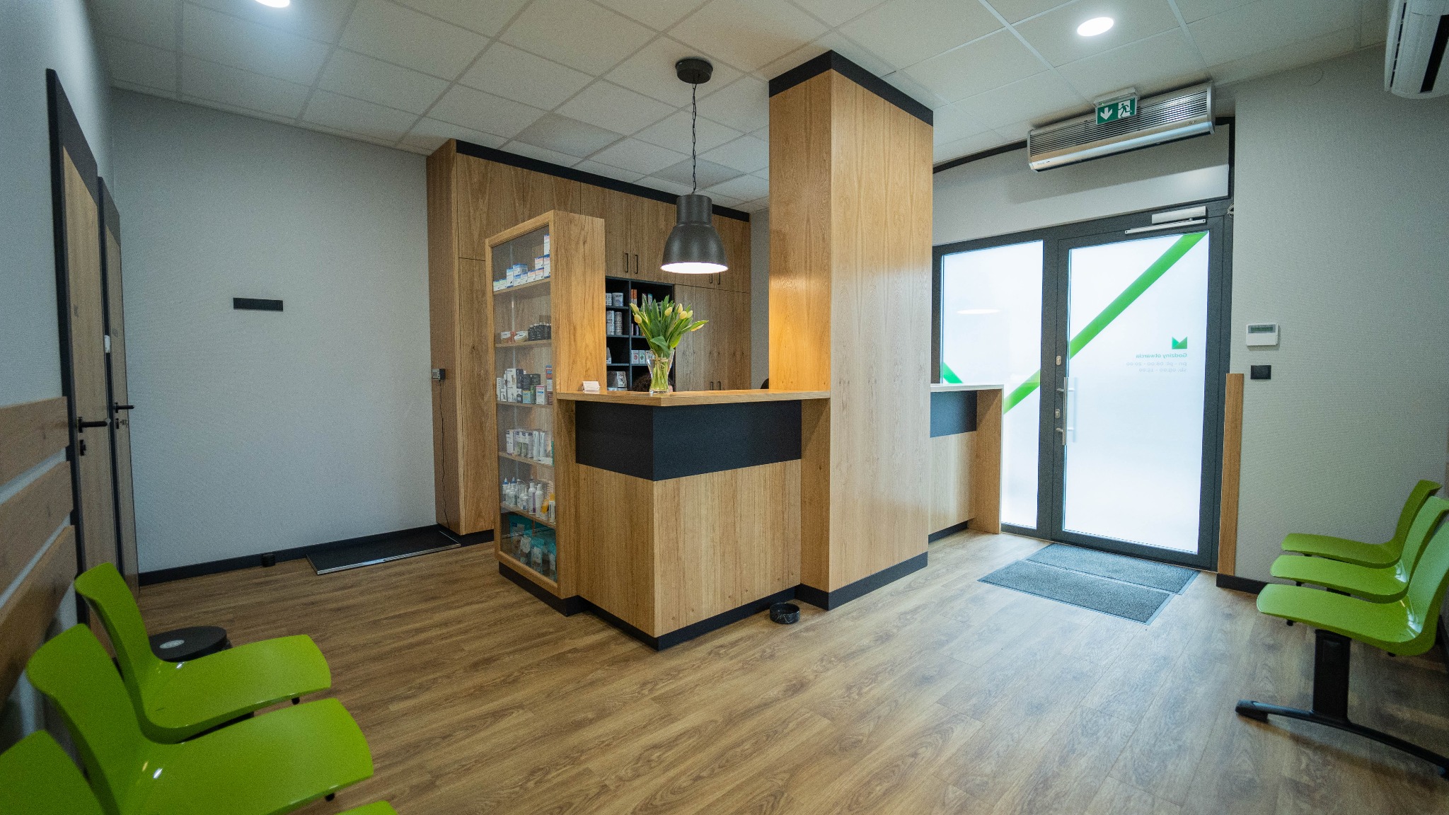 Jasne wnętrze recepcji z drewnianymi elementami i zielonymi krzesłami dla klientów, widoczne kwiaty w wazonie na ladzie.