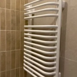 Biały, designerski grzejnik łazienkowy na tle beżowych płytek. Widok z perspektywy narożnika łazienki. Minimalistyczny design, nowoczesne wykończenie.