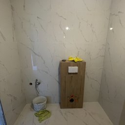 GRbud György Román - Wnętrze łazienki w trakcie prac: białe płytki imitujące marmur na ścianach i podłodze, zabudowa stelaża WC z widocznymi przyłączami, wiadro i ściereczka na podłodze.