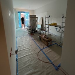 GRbud György Román - Wnętrze w trakcie malowania: zabezpieczona podłoga, drabina, puszki z farbą, okno osłonięte folią, widoczny fragment pomalowanej ściany i pracownik przy oknie.