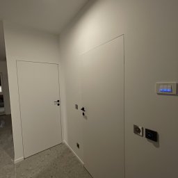 GRbud György Román - Minimalistyczny korytarz z dwiema białymi, ukrytymi drzwiami i szarą posadzką lastryko. Na ścianie włączniki z niebieskim podświetleniem i gniazdka.