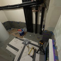 GRbud György Román - Remont łazienki w trakcie: ułożone płytki podłogowe i ścienne z użyciem klipsów poziomujących, widoczne rury instalacji, narzędzia i laser krzyżowy na statywie.
