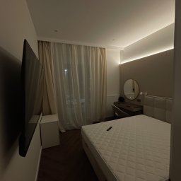 GRbud György Román - Nowoczesna sypialnia z dużym telewizorem na ścianie, łóżkiem z pikowanym zagłówkiem i toaletką z okrągłym lustrem, oświetlona listwą LED.