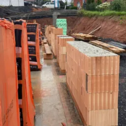 Wznoszenie muru z bloczków ceramicznych, widoczne opakowania bloczków w pomarańczowej folii, wiadro z zaprawą i kielnia, etap budowy domu.