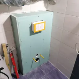 Konstrukcja z płyty kartonowo-gipsowej zabudowująca stelaż WC w trakcie montażu, widoczne otwory na przyłącza, poziomica i folia ochronna na ścianie.