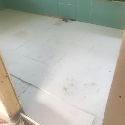 GRbud György Román - Pokój w trakcie prac wykończeniowych: ściany pokryte zielonymi płytami, podłoga wyłożona białymi płytami styropianowymi, widoczne okno z preparatami na parapecie.