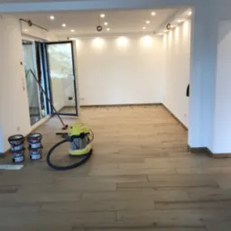 Jasne, drewnopodobne płytki podłogowe ułożone w nowym pomieszczeniu, widoczne pojemniki z klejem, odkurzacz przemysłowy oraz krzyżyki dystansowe, otwarte okno balkonowe.