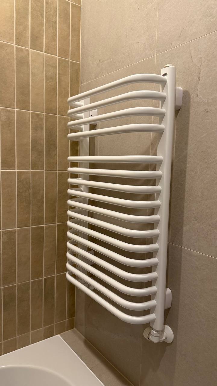 Biały, designerski grzejnik łazienkowy na tle beżowych płytek. Widok z perspektywy narożnika łazienki. Minimalistyczny design, nowoczesne wykończenie.